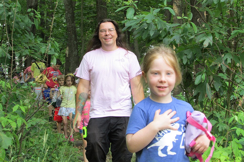 a line of children and an adult leader on a hike in the woods, walking towards camera